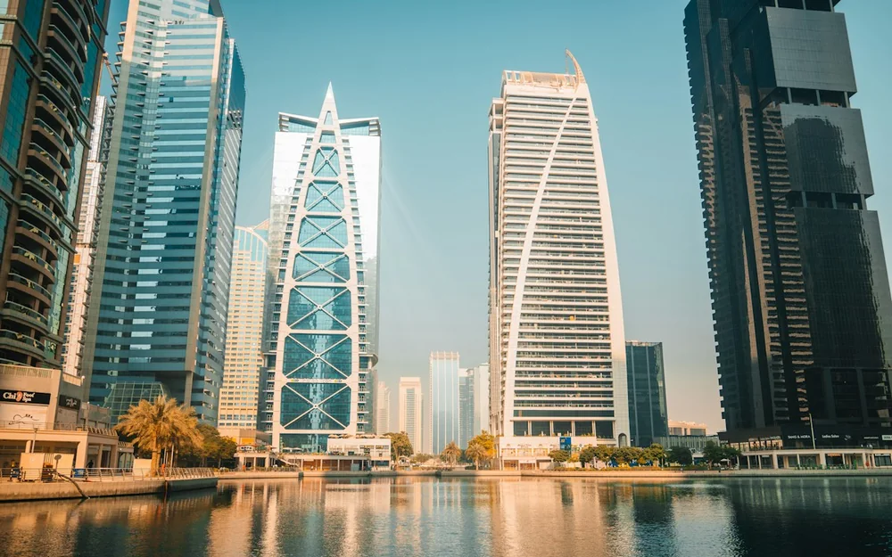 Dubai towers reflected in calm waterfront water beneath a blue sky.